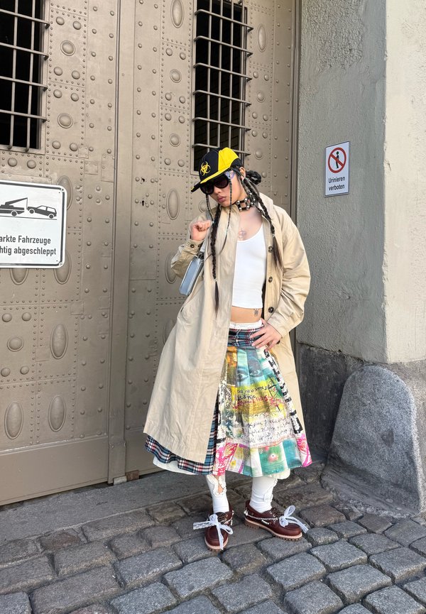 Persona con indosso un cappotto beige, una gonna patchwork colorata, una maglietta bianca e un berretto giallo-nero, in piedi su una strada lastricata vicino a una grande porta metallica con cartelli in tedesco.