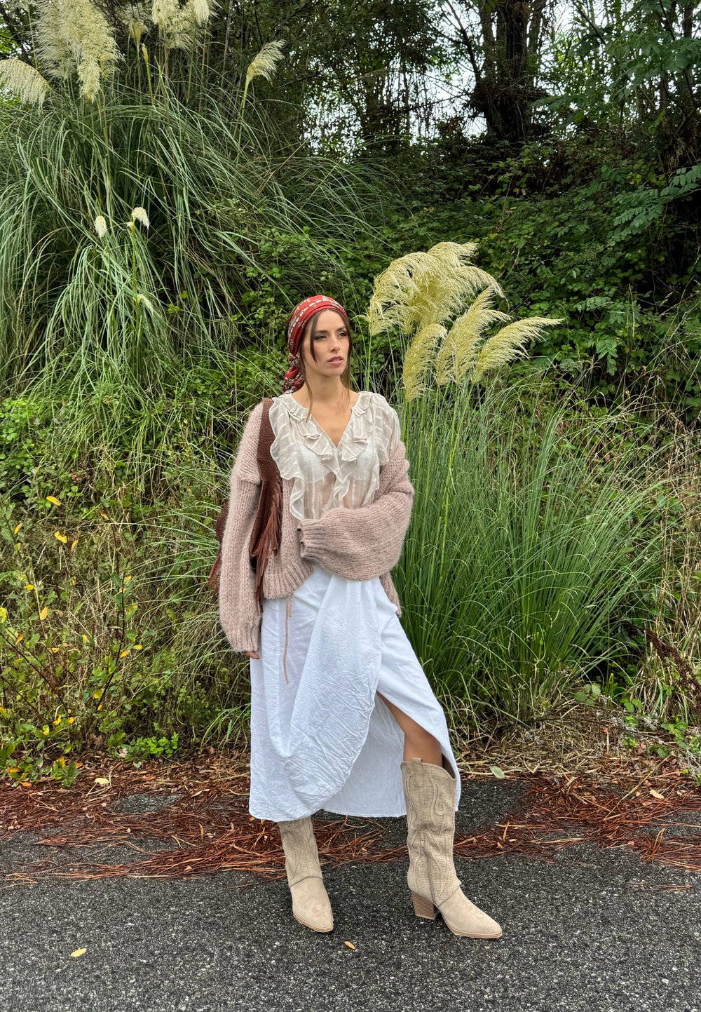 Mujer con una blusa de volantes en beige claro, un cárdigan rosa de punto oversized, una falda blanca con una abertura lateral y botas de caña alta en beige claro.