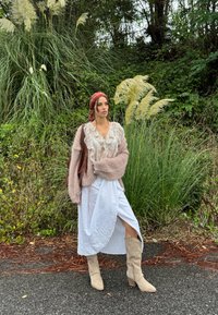 Mujer con una blusa de volantes en beige claro, un cárdigan rosa de punto oversized, una falda blanca con una abertura lateral y botas de caña alta en beige claro.