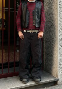 Gilet en cuir noir sur une chemise à manches longues rouge et noire à rayures, associé à un pantalon noir ample et une boucle de ceinture en forme d'étoile en argent.