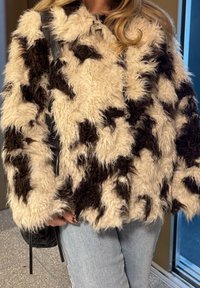 Műszőrméből készült kabát, krémszínű és barna tehénmintával, shaggy, bolyhos textúrával és enyhén túlméretezett szabással, kék farmerrel párosítva.