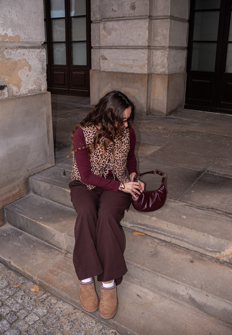 Persona, kas sēž uz akmens kāpnēm, ģērbusies leopardu raksta vestē virs bordo krekla, brūnās platās biksēs un gaišās kurpēs, tur rokā bordo somu.
