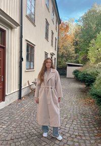 Abrigo suave de un beige claro con un amplio collar y cinturón, usado sobre una blusa clara y vaqueros acampanados azules. El conjunto se complementa con zapatillas blancas.