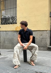 Vállalkozó stílusú öltözet fekete galléros pólóval, világosszürke terepmintás nadrággal és papucsokkal, egy fekete oszlopon ülve, egy texturált fal előtt.