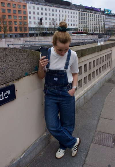 Pantalones de mezclilla azul con un corte relajado, camiseta blanca debajo, sosteniendo una taza de café negra; zapatillas con tres franjas en colores contrastantes.