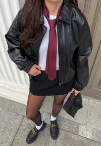 Sort faux læderjakke med ribbede manchetter, hvid t-shirt, burgunder slips, sort mini nederdel, gennemsigtige sorte strømpebukser, og sorte loafers med guld accenter.