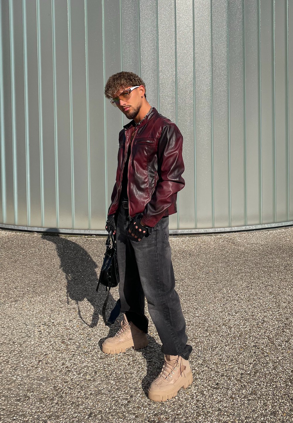 Un homme portant une veste en cuir bordeaux, des gants noirs, des lunettes de soleil, un jean noir et des bottes à plateforme beiges se tient sur du gravier près d'un mur en verre givré.