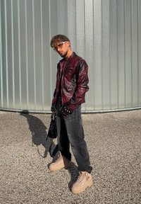 Un homme portant une veste en cuir bordeaux, des gants noirs, des lunettes de soleil, un jean noir et des bottes à plateforme beiges se tient sur du gravier près d'un mur en verre givré.