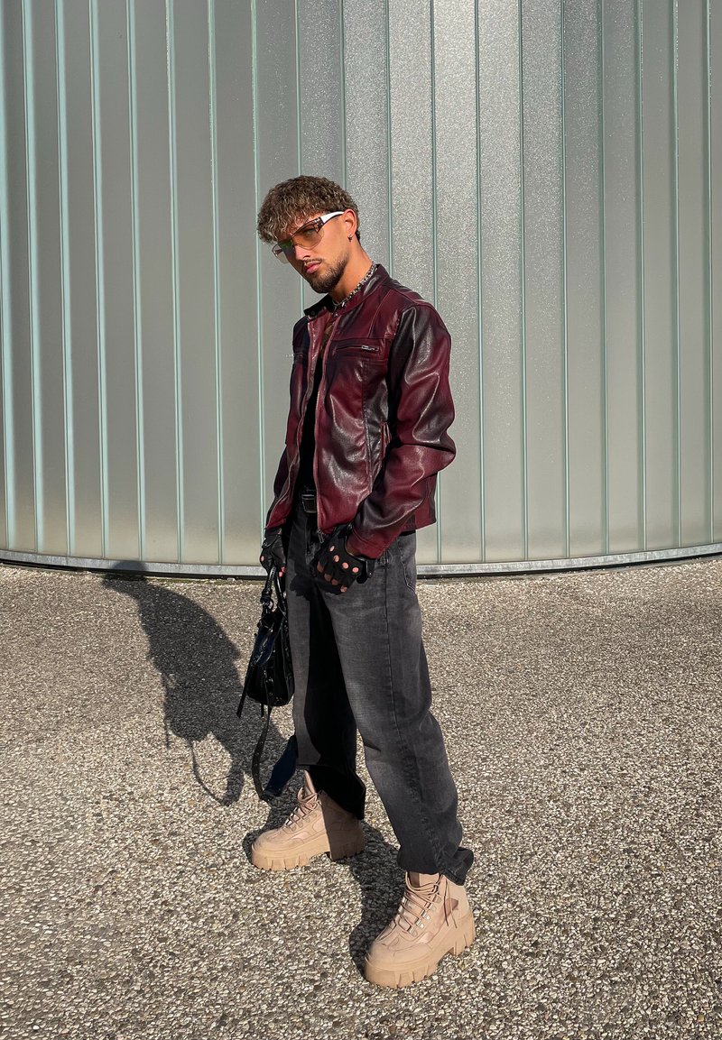 A man dressed in a burgundy leather jacket, black gloves, sunglasses, black jeans, and beige platform boots is standing on gravel next to a frosted glass wall.