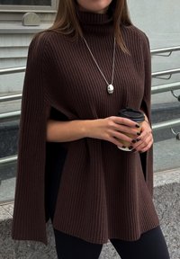 Poncho marrone a coste con colletto alto, dotato di aperture laterali e vestibilità rilassata. Tiene in mano una tazza di caffè nera, abbinato a leggins neri.