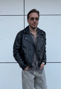 Zwarte leren motorjack met zilveren hardware, voorzien van ritszakken en een notched kraag, gecombineerd met een grijs shirt en lichte jeans.