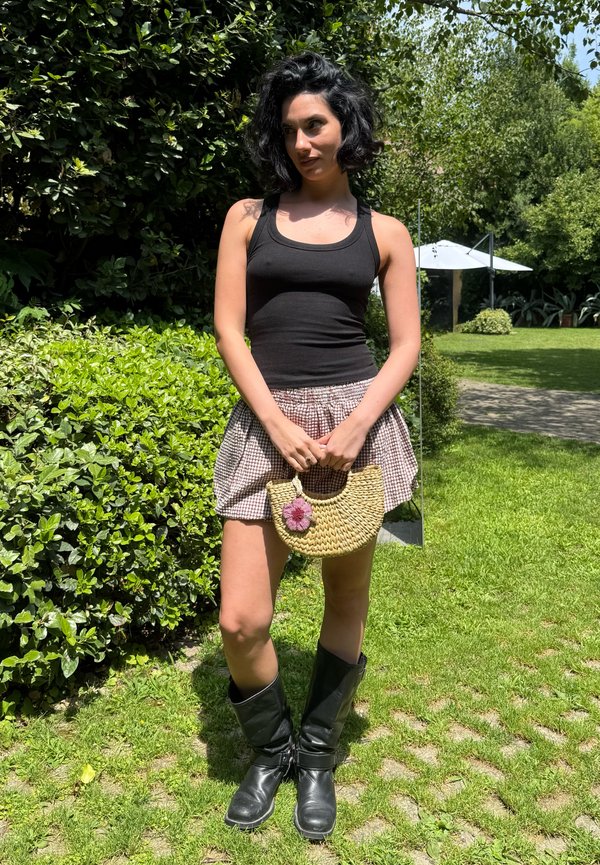 Frau mit kurzen lockigen Haaren, die ein schwarzes Tanktop, karierte Shorts, schwarze Stiefel trägt und eine Strohtasche mit einer lila Blume hält, steht auf Gras.