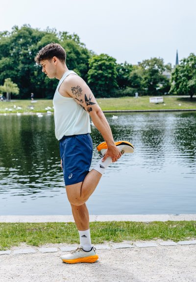Hombre atlético con camiseta sin mangas de color azul claro y pantalones cortos navy estira la pierna junto al agua. Lleva zapatillas de correr blancas y naranjas con calcetas blancas.