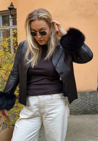 Giacca nera in pelle con rifinitura in pelliccia sulle maniche, indossata sopra una maglia marrone e pantaloni bianchi. Accessori comprendono occhiali da sole tondi e una borsa.