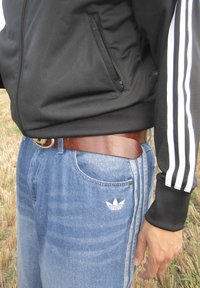 Fekete cipzáras dzseki, három fehér csíkkal a hónalján, kék farmernadrággal párosítva, amin fehér Adidas logó és barna bőr öv található.