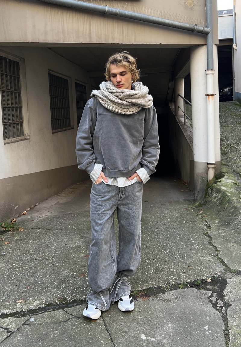 Grey sweatshirt with ribbed cuffs, layered over a white striped shirt. Loose grey jeans and white trainers with black accents. Textured knit scarf.