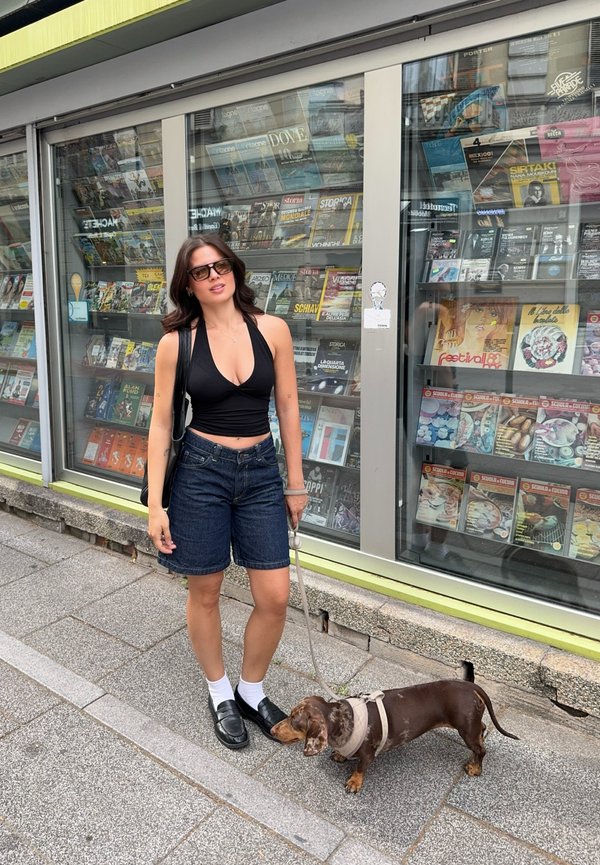 Femme portant un haut noir et un short en jean, debout sur le trottoir, tenant la laisse d'un petit chien marron devant la vitrine d'un magasin de magazines.