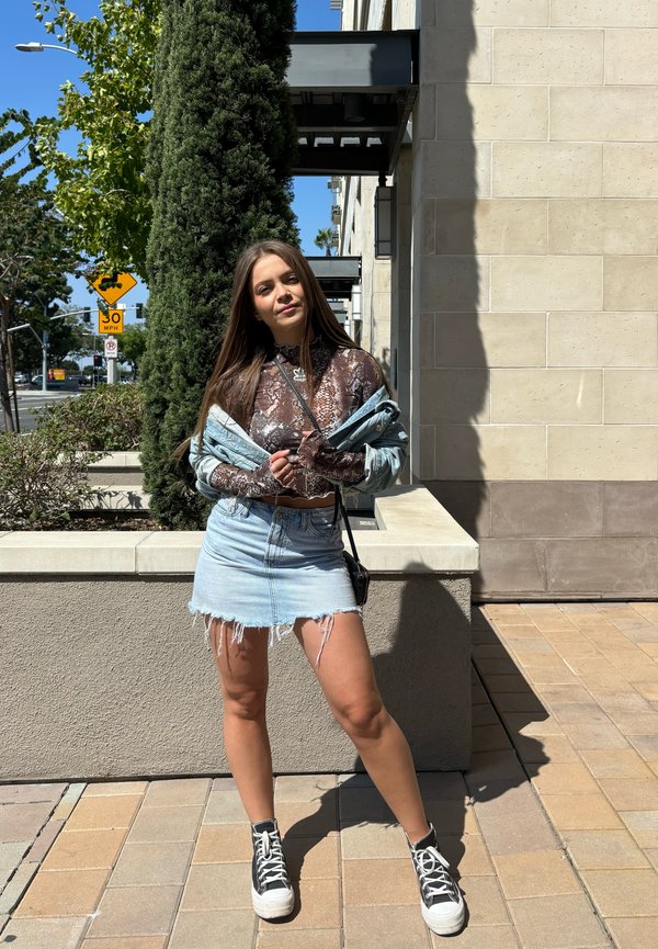 Falda de mezclilla con dobladillo deshilachado, combinada con una blusa transparente marrón y blanca de estampado, superpuesta con una chaqueta de mezclilla. Zapatillas altas negras.