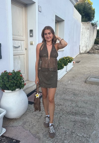 Joven mujer con un vestido halter marrón y transparente con patrones de estrellas, sosteniendo un bolso marrón con flecos y una flor blanca, de pie en un camino de piedra cerca de edificios blancos.