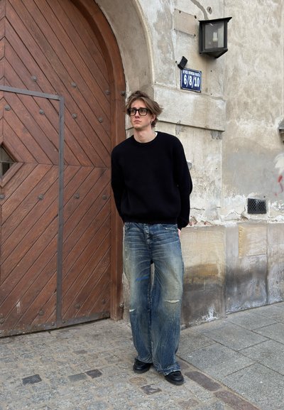 Joven con gafas, suéter negro y vaqueros anchos, junto a una gran puerta arqueada de madera en la fachada envejecida de un edificio de piedra.
