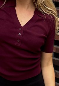 Burgundy short-sleeve top with a V-neck, featuring a buttoned placket and ribbed texture. The fabric appears smooth and fitted.