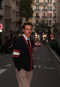 Strikket marineblå cardigan med røde og hvite striper, kombinert med en lyseblå skjorte og mørkt slips, stående i en bygate i skumringen.