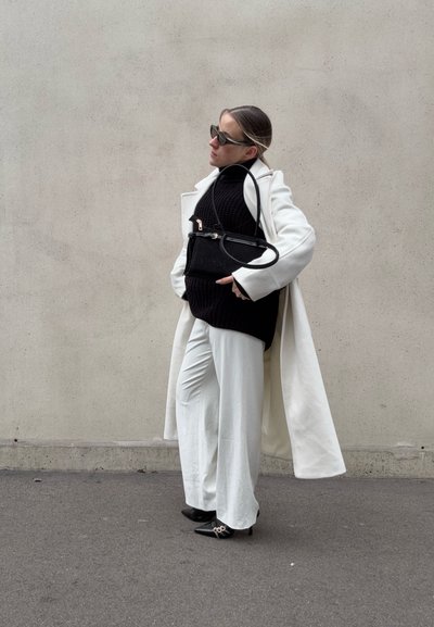 Mujer con abrigo blanco y pantalones, cuello alto negro y tacones, sosteniendo bolso negro, de pie frente a una pared beige lisa.