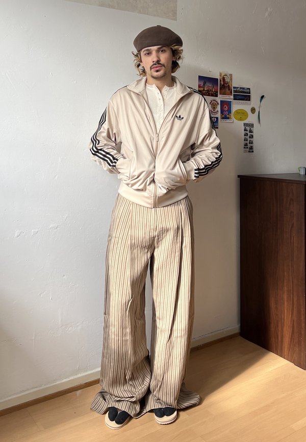 Joven con boina marrón, chaqueta beige de Adidas, pantalones anchos beige con rayas y sandalias negras, de pie dentro de un edificio cerca de una pared con pequeños carteles.