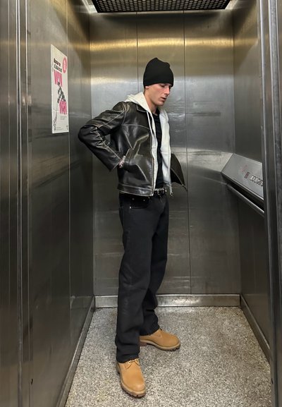 Chaqueta de cuero negra con una sudadera gris debajo, jeans negros y botas beige en un ascensor metálico con un cartel en la pared.