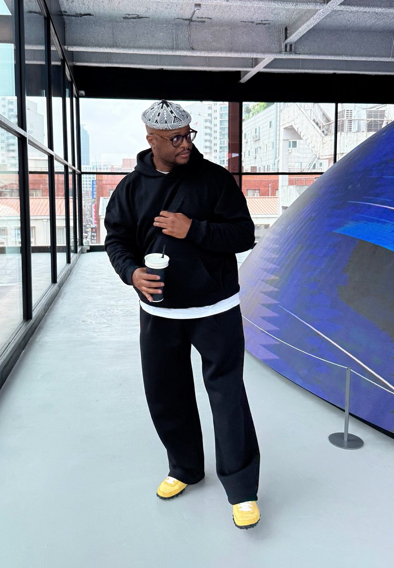 Homme en sweat à capuche noir, casquette à motifs et baskets jaunes tenant une tasse, debout à l'intérieur près d'une grande sculpture géométrique bleue.