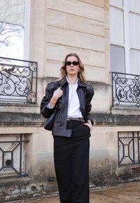 Black leather jacket with a structured design, worn over a white shirt and black skirt. Accessories include narrow sunglasses and a belt.