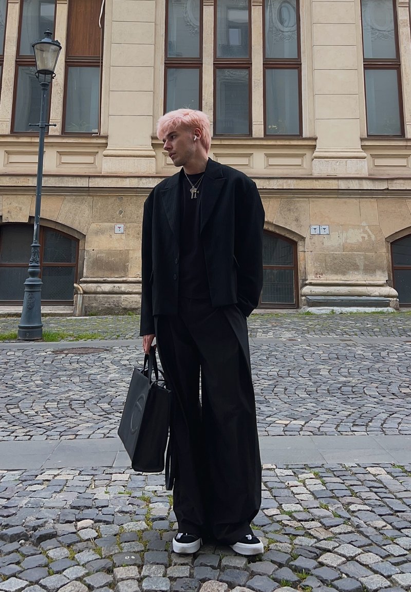 Jeune personne aux cheveux roses portant des vêtements entièrement noirs et tenant un sac noir, se tient dans une rue pavée devant un bâtiment beige.