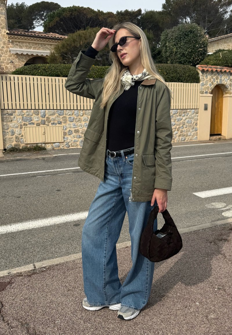 Woman wearing sunglasses, a green jacket, black top, wide jeans, and white trainers, holding a black handbag, standing on a street with stone buildings behind her.