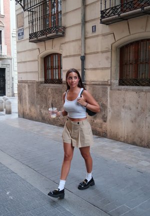 Una mujer con una camiseta sin mangas gris y una falda beige camina por una calle de piedra, sosteniendo una bebida, llevando zapatos negros y calcetines blancos.