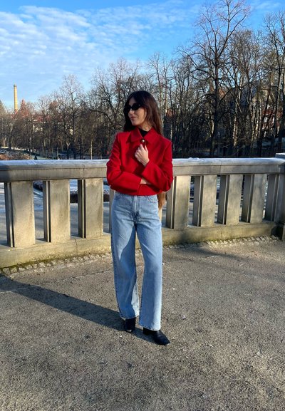 Cazadora roja cortada, vaqueros de tiro alto y pernera ancha de color azul claro, y botas negras de tacón puntiagudo, de pie en un camino de piedra cerca de una barandilla.