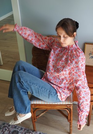 Blusa floral con cuello fruncido, patrones rojos y morados, mangas largas y voluminosas; combinada con jeans de campana azules y zapatos blancos.
