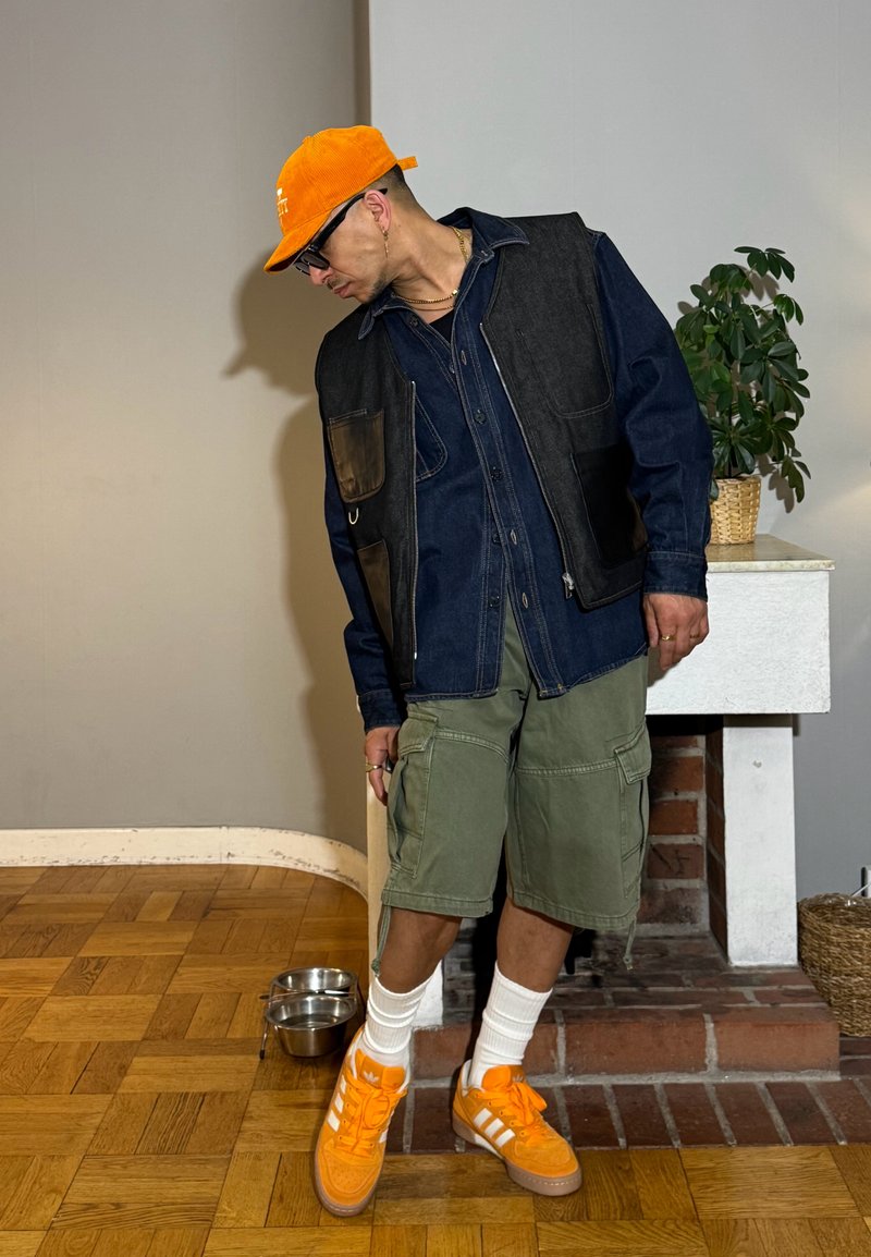 Homme portant une casquette orange et des baskets, une veste marine et un short cargo vert, debout à l'intérieur près d'une plante et des gamelles pour animaux.