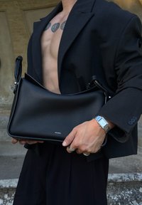Person, der ein offenes schwarzes Sakko trägt und Brust-Tattoos zeigt, hält eine strukturierte schwarze Pedro-Schultertasche und trägt eine silberne Uhr sowie Ringe.