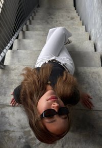 En person med långt brunt hår bär svarta solglasögon och en svart topp, lutad mot gråa betongtrappor, med vita byxor och svart bälte.