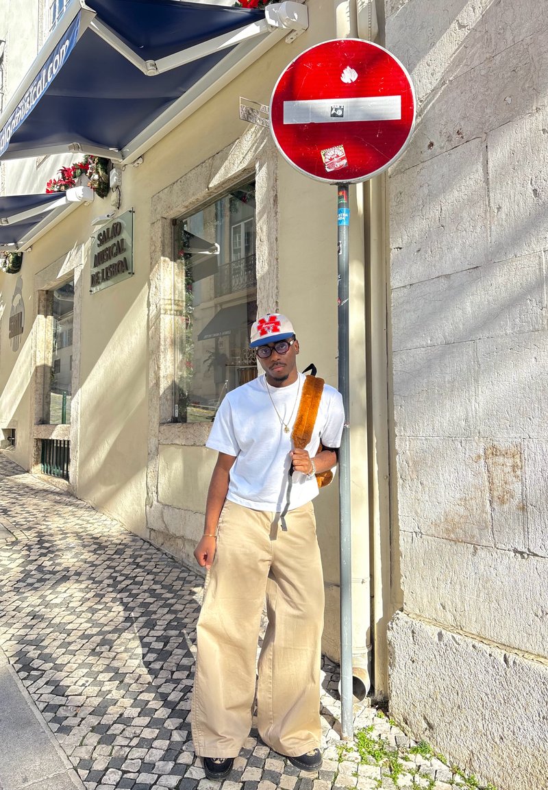 Une personne portant une chemise blanche et un pantalon beige à jambes larges se tient à côté d'un panneau rouge d'interdiction d'entrée contre un mur en pierre texturé avec un trottoir ombragé.