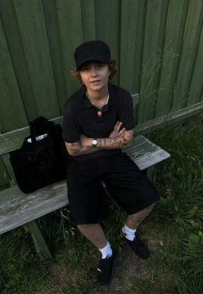 Gorra negra, camiseta polo negra y pantalones cortos. Joyería de plata, incluyendo un colgante en forma de corazón, llevada en brazos cubiertos de tatuajes. Zapatillas negras y bolsa.