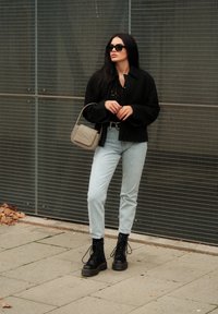 Μαύρο μπουφάν, μαύρη μπλούζα με λεπτομέρειες, ανοιχτό μπλε τζιν, μαύρες στρατιωτικές μπότες με κορδόνια και γκρίζα τσάντα χιαστί σε μεταλλικό φόντο.