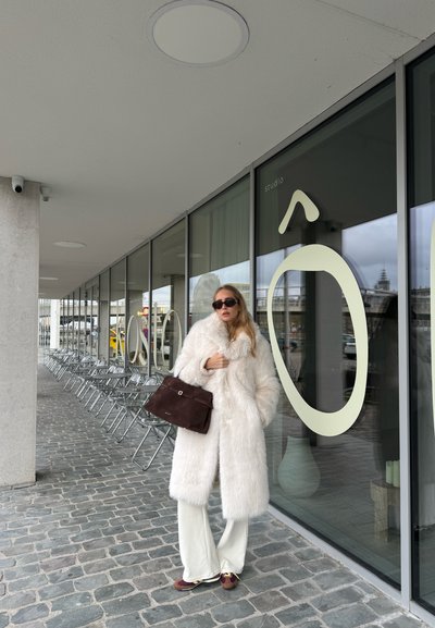 Abrigo de piel sintética blanco, pantalones de pierna ancha color beige claro, bolso marrón oscuro, gafas de sol negras de gran tamaño. Pavimento de piedra lisa y escaparate de cristal.