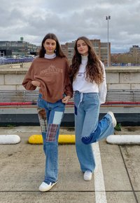 To personer står side om side. Den ene har en brun sweatshirt på med tekst og lapper og slidte jeans; den anden har en hvid bluse og blå jeans.