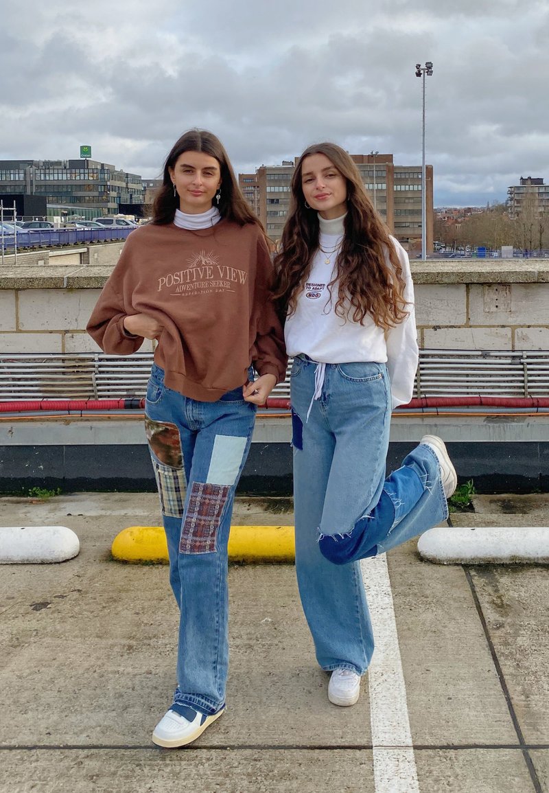 To personer står side om side. Den ene har en brun sweatshirt på med tekst og lapper og slidte jeans; den anden har en hvid bluse og blå jeans.