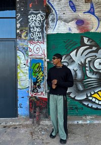 Černý svetr, zelené a černé rozdělené kalhoty a sluneční brýle před barevnou graffiti zdí. Rysy zahrnují různé textury a odvážné vzory.