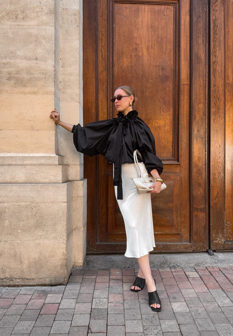 Black puff-sleeve blouse with ruffle neck, white satin skirt, black heeled sandals, holding a small white handbag against a wooden backdrop.