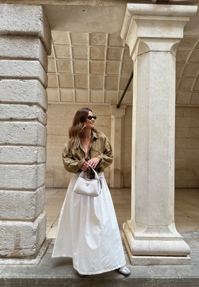 Camisa oversize verde oliva, falda larga blanca, zapatos gris claro, bolso blanco con logo, con columnas de piedra texturizadas de fondo.