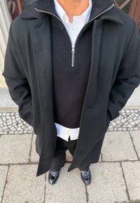 Homme debout en plein air, portant un pardessus noir, un pull à fermeture éclair noir, une chemise blanche boutonnée, un pantalon noir et des chaussures en cuir noires.