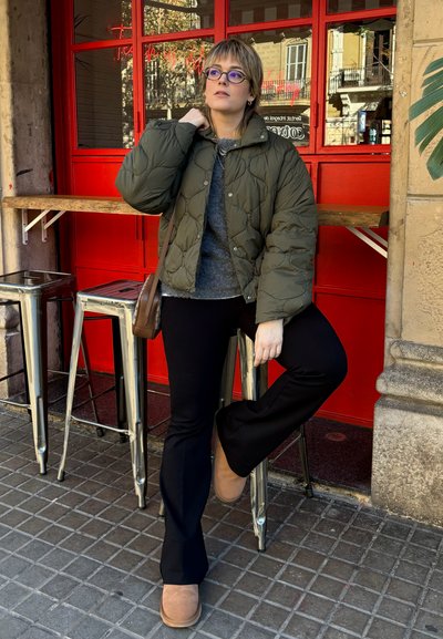 Chaqueta acolchada verde oliva, suéter gris debajo, pantalones de campana negros, botas de tobillo marrones. El fondo presenta una pared roja y taburetes de metal.